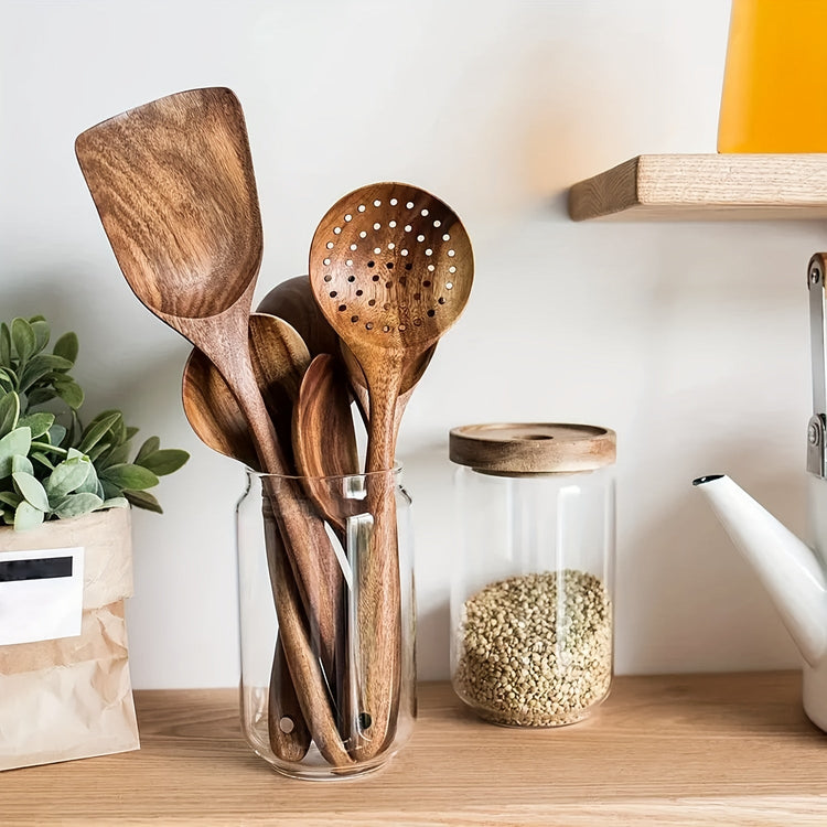 Set of wooden utensils in a glass jar on a kitchen counter with a plant and jar of seeds in the background.