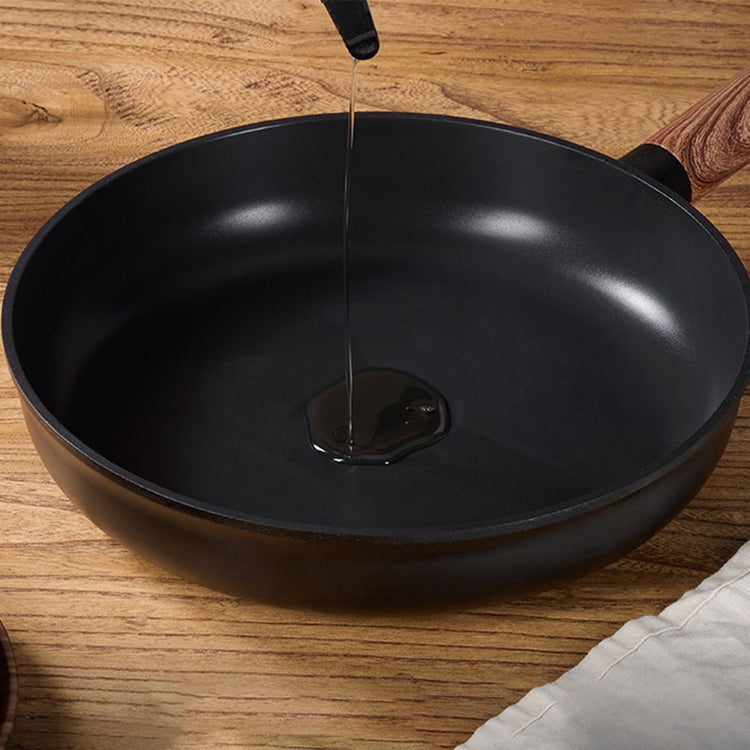 Black frying pan on a wooden surface with oil being poured into it.