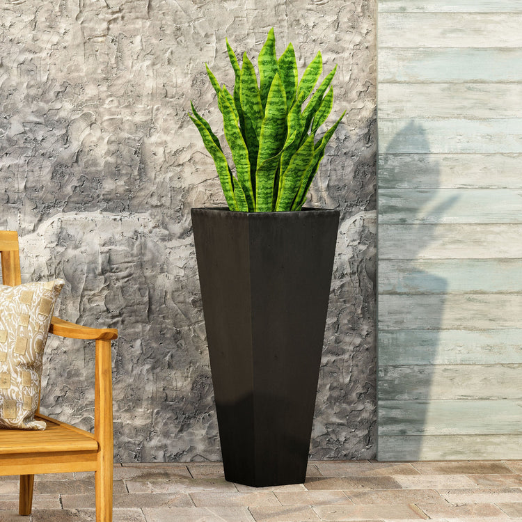 Tall black planter with a green plant against a stone wall.
