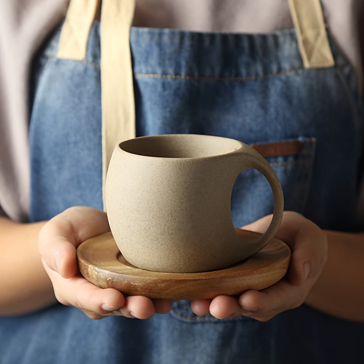 Person holding a beige ceramic cup on a wooden saucer, wearing a blue apron.