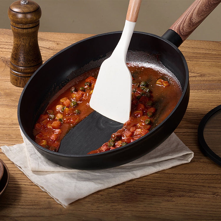 Black frying pan with tomato-based dish and white spatula on a wooden table