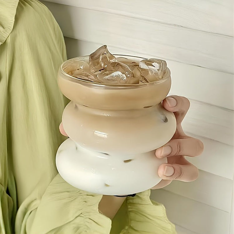 Person holding a glass with a creamy beverage and ice cubes against a neutral background