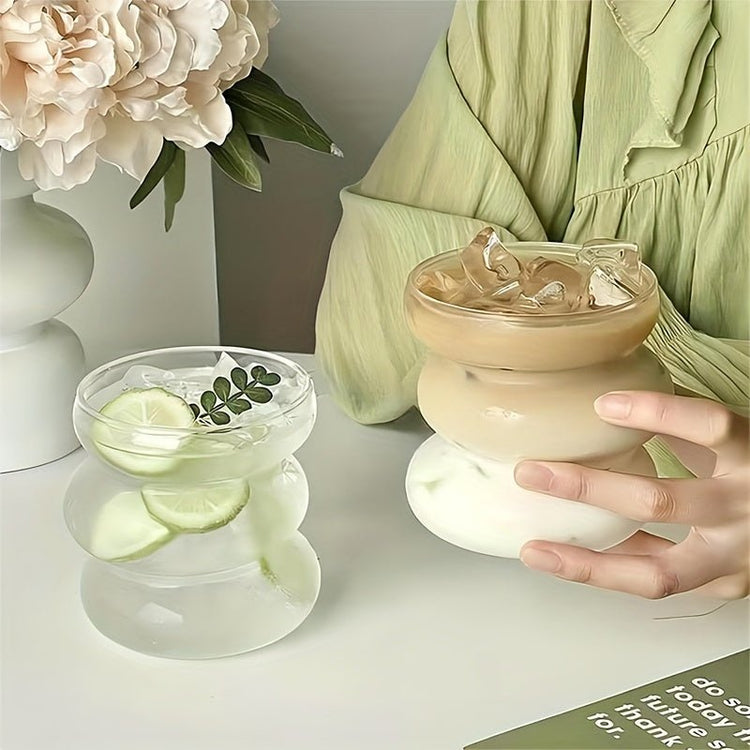 Person holding a glass with a creamy beverage and ice cubes, next to a small bowl with lime slices and mint leaves on a light surface.