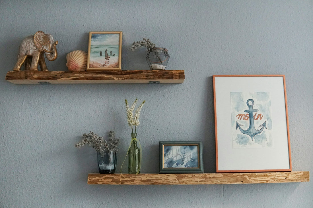 Rustic floating wooden shelves on a blue-grey wall displaying a brass elephant figurine, seashell, watercolor prints, dried flowers in glass vases, and a framed anchor illustration