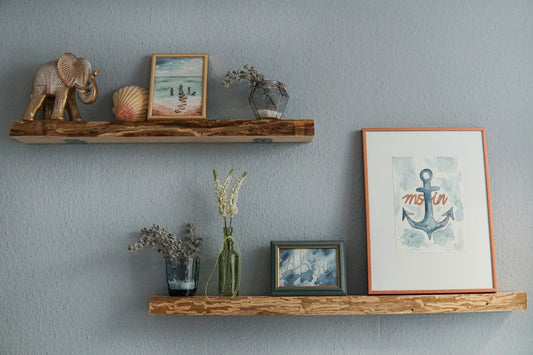 Rustic floating wooden shelves on a blue-grey wall displaying a brass elephant figurine, seashell, watercolor prints, dried flowers in glass vases, and a framed anchor illustration