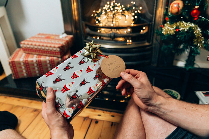 Hands holding a Secret Santa gift with festive wrapping paper by a log burner with fairy lights