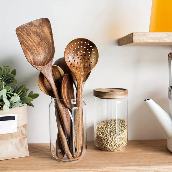 Set of wooden utensils in a glass jar on a kitchen counter with a plant and jar of seeds in the background.