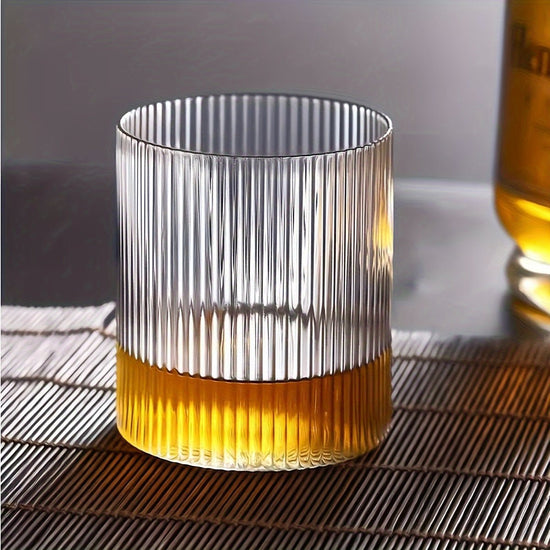 Clear glass tumbler with ribbed design filled with a yellowish liquid on a textured surface.