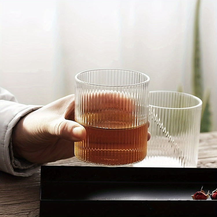 Hand holding a glass of amber liquid with another glass in the background on a wooden surface.