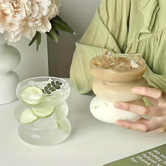 Person holding a glass with a creamy beverage and ice cubes, next to a small bowl with lime slices and mint leaves on a light surface.