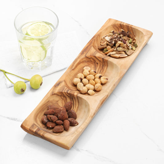 Wooden snack tray with almonds, peanuts, and pistachios on a white surface with a glass of water and lime.