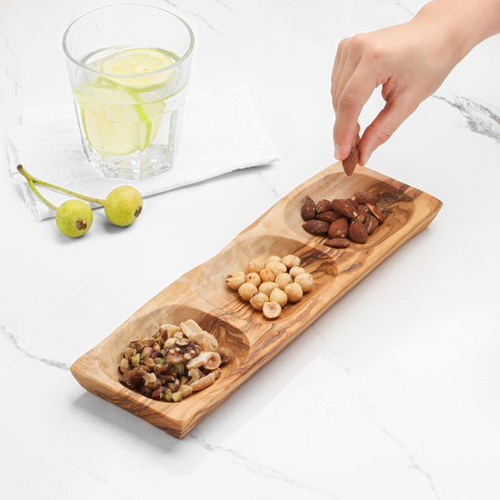 Wooden platter with assorted nuts on a white surface, with a hand reaching in to pick up a nut.